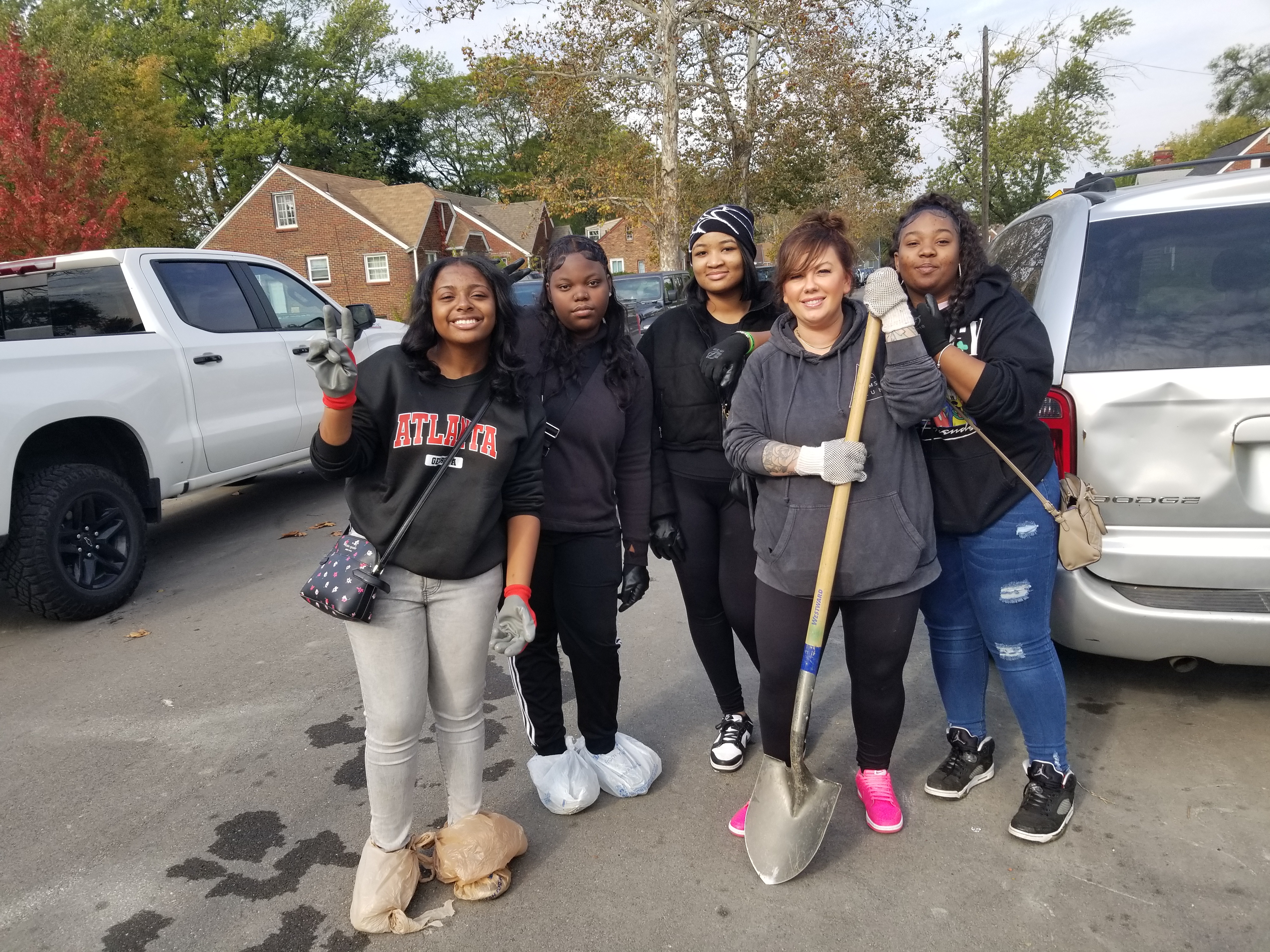 Five young people stand, smiling and making peace signs on Kensington Ave in the autumn sun. One person holds a shovel, and a few have their shoes covered so they can work in dirt.