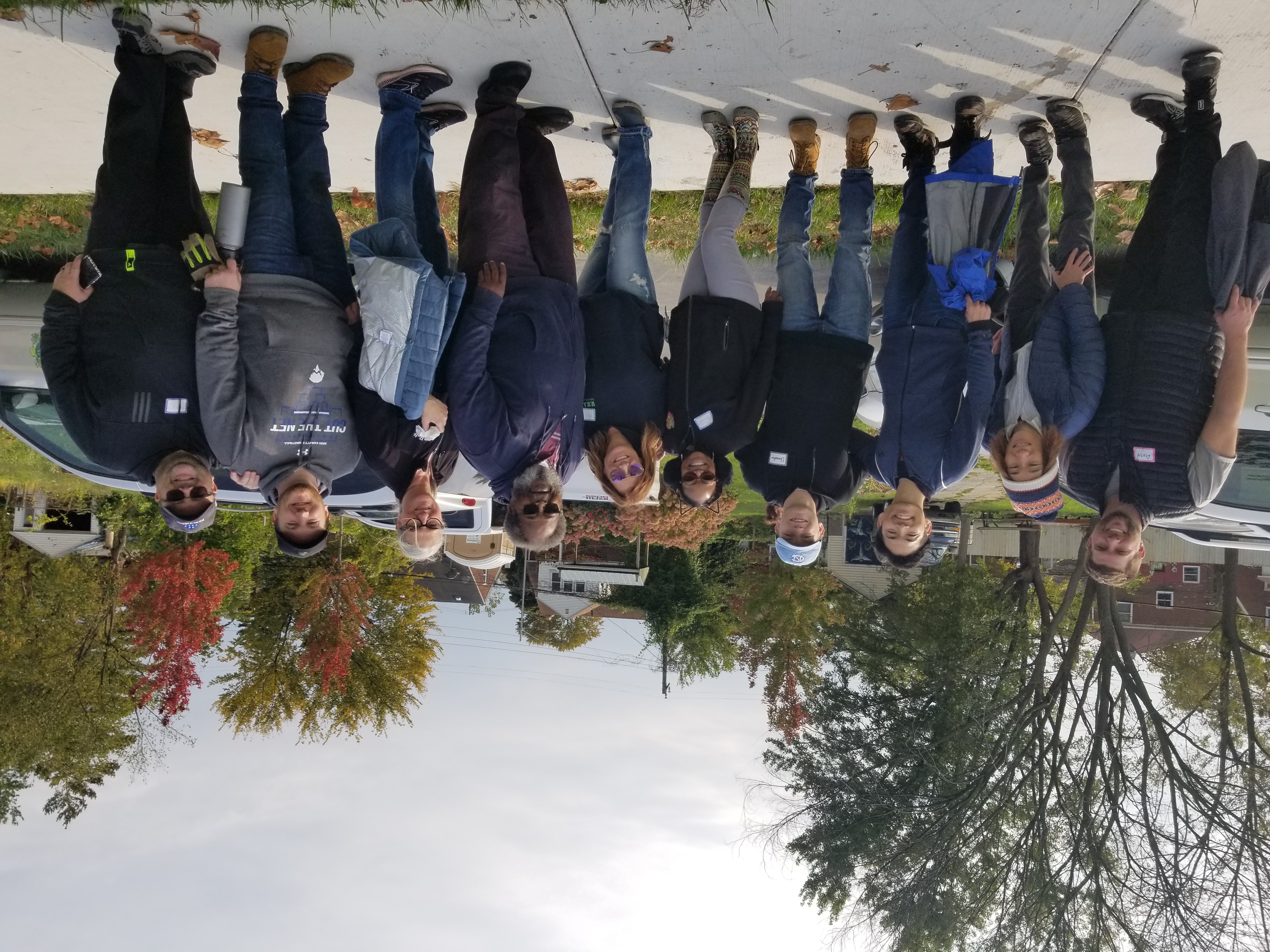 Ten people with nametags, including Mose Primus, smile at the camera on a crisp autumn day. They stand on the sidewalk in front of the 4 Angels Community Garden area.