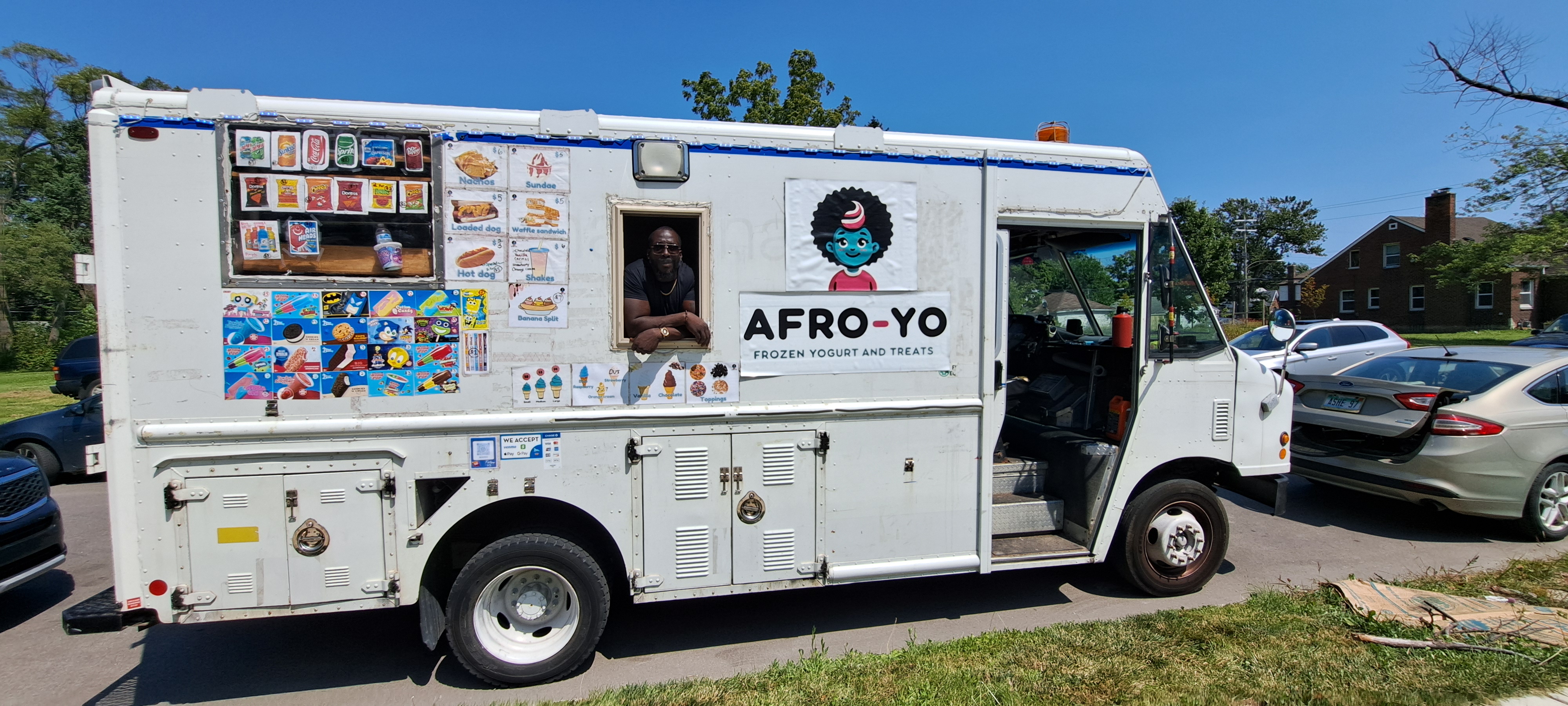 Afro-yo vendor standing at the window of a food truck decorated with brightly colored menu items.