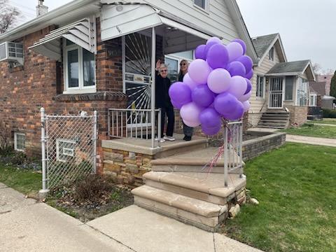 From the front porch of a brick house decorated with a bunch of purple balloons, Ms. Lottie waves to the camera.