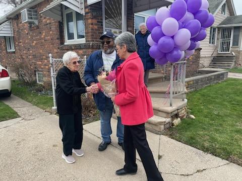 In front of a house decorated with a bunch of purple balloons, Rep. Paiz shakes hands with Ms. Lottie and presents her a bouquet of flowers.