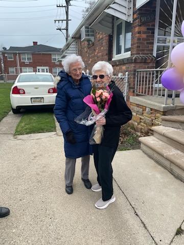 Ms. Lottie poses with a friend on the front walk of her home. She smiles, holding a bouquet of flowers.