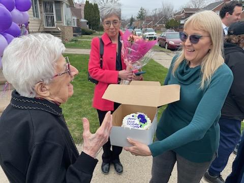 Ms. Lottie smiles as she is presented with a birthday cake. Nearby, Rep. Veronica Paiz waits with a bouquet of flowers.