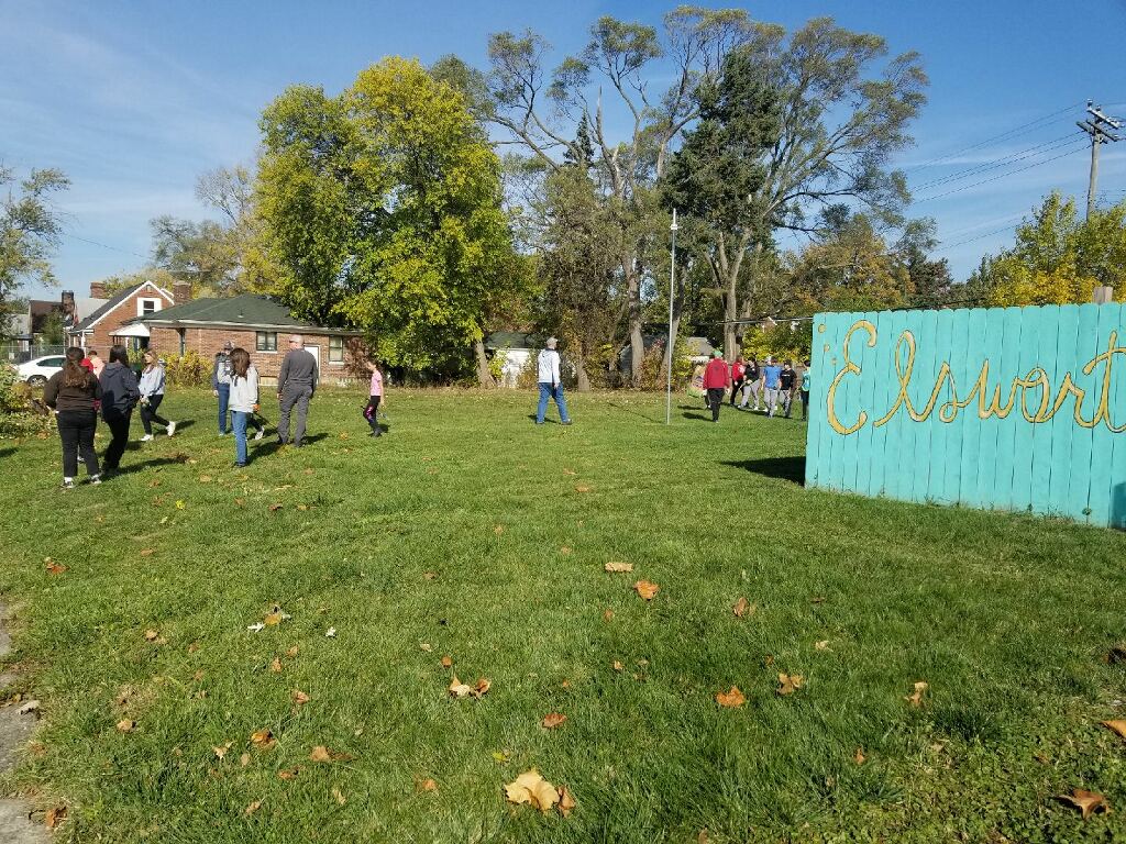 People scattered about a field in the early autumn sunshine, near a wooden fence painted a playful blue. On the fence, yellow calligraphy reads, "Elsworth."