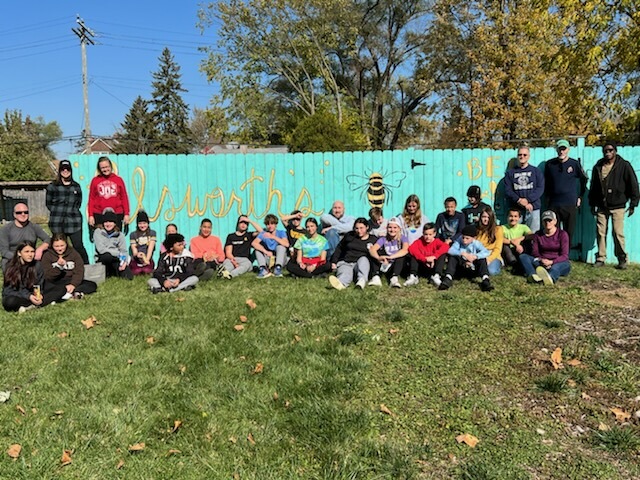 A group of maybe 20 people, mostly white, pose against a light blue fence with yellow calligraphy reading "Ellsworth's bee adventure."