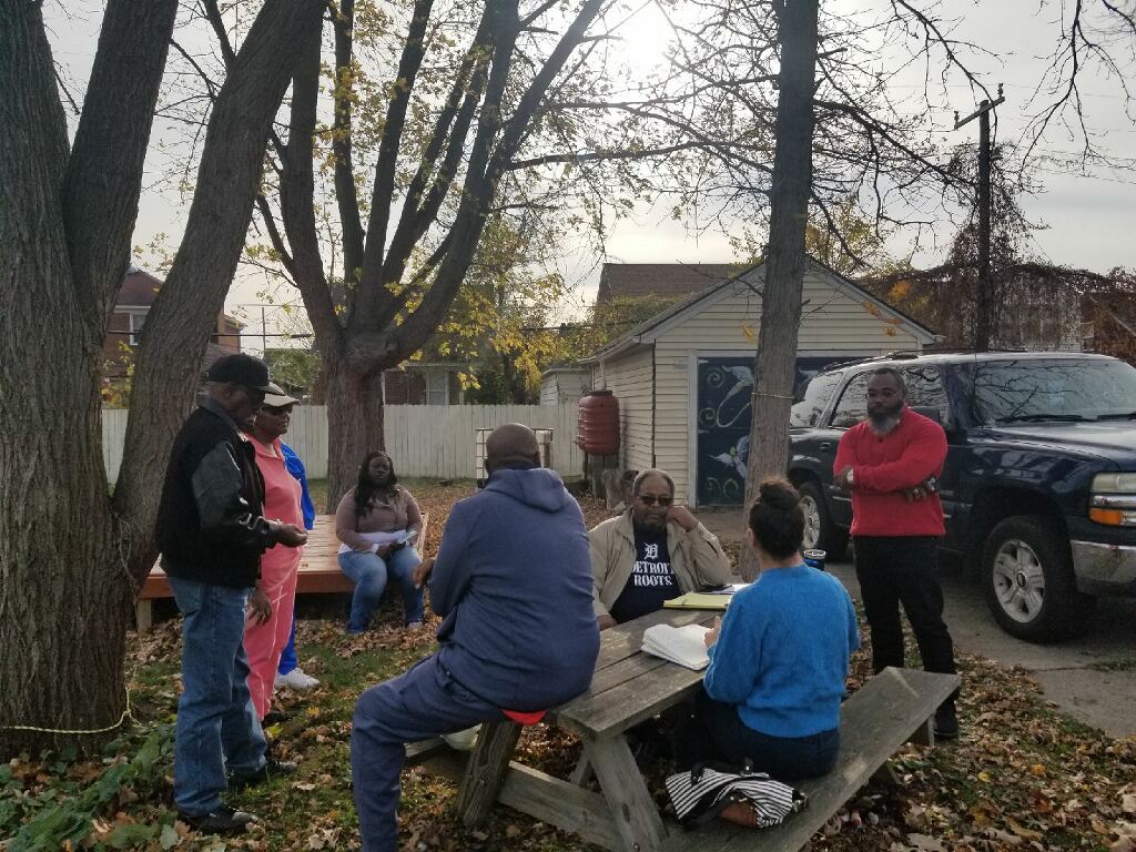 Eight people gather around a picnic table in a park, while Mose speaks animatedly with another attendee.